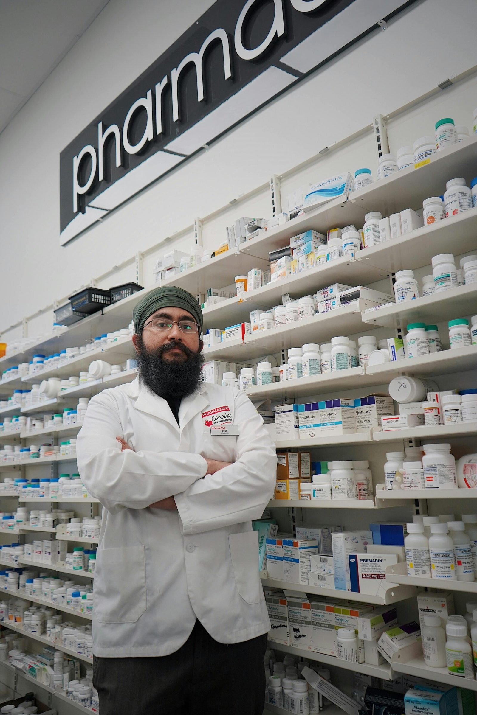 Pharmacist with turban and glasses in a pharmacy, standing in front of medicine shelves.